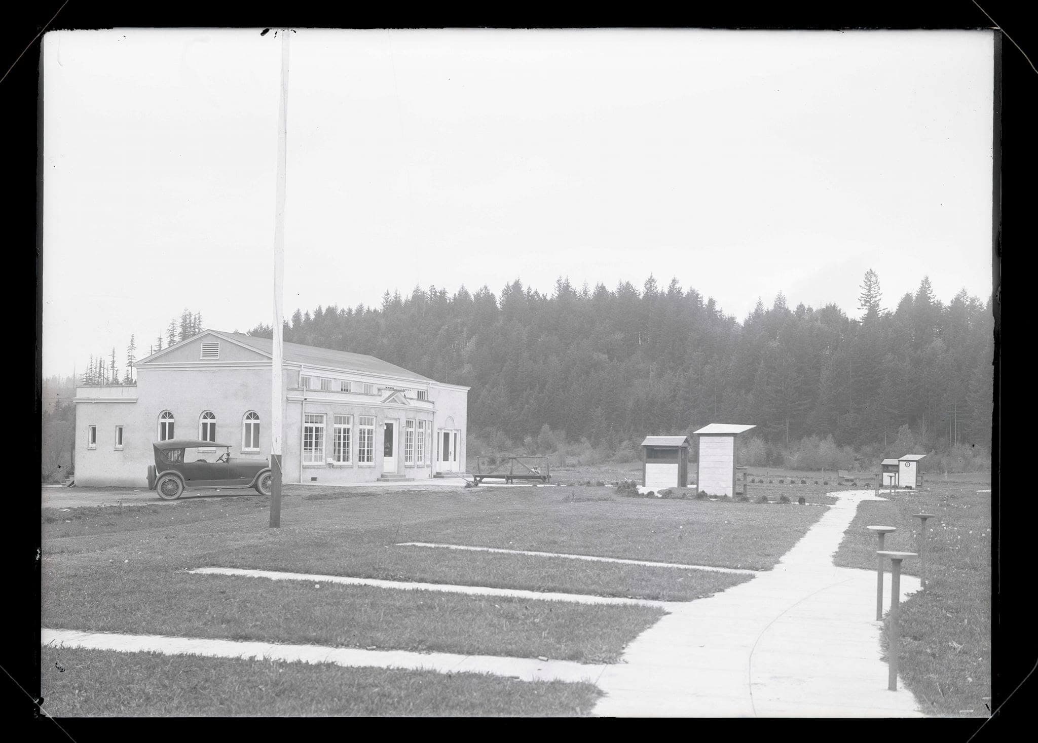 A white clubhouse with a flagpole and walkways, an early automobile parked outside